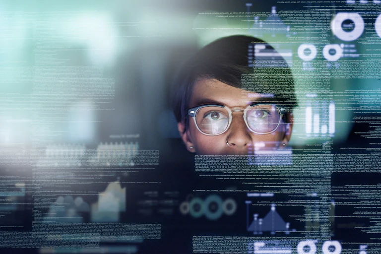 A person with short dark hair, glasses, and a nose ring looks at a futuristic screen with writing and charts.