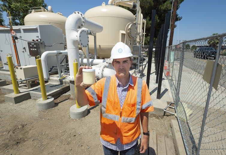 A man in a reflective vest holds a jar in front of water equipment.