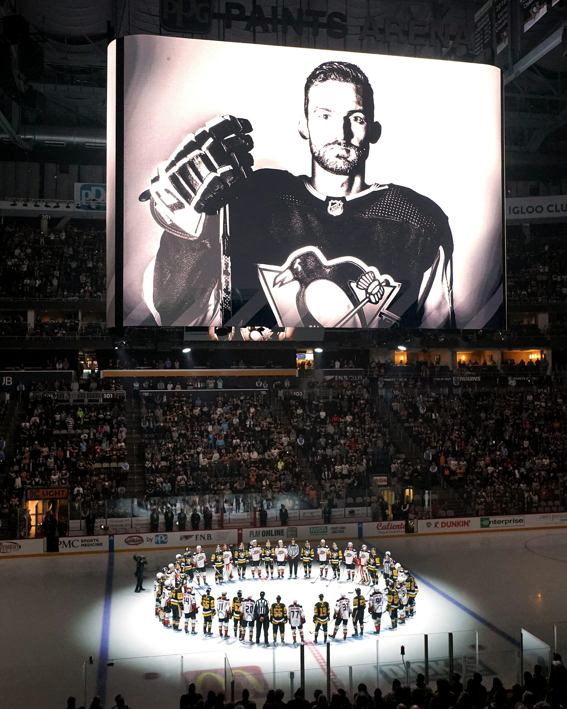 A black-and-white photo of a hockey player is displayed on a large screen above an ice hockey rink. A circle of hockey players stand on the ice below it, illuminated by a spotlight.