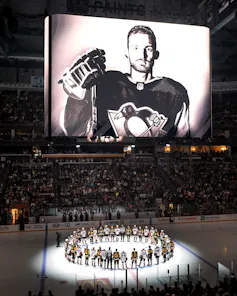 A black-and-white photo of a hockey player is displayed on a large screen above an ice hockey rink. A circle of hockey players stand on the ice below it, illuminated by a spotlight.