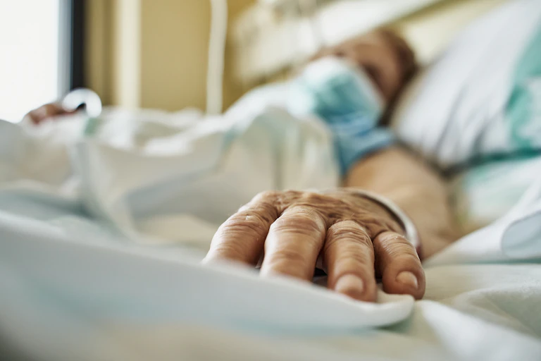 Paciente usando máscara deitado em uma cama de hospital, com a mão estendida em direção à câmera