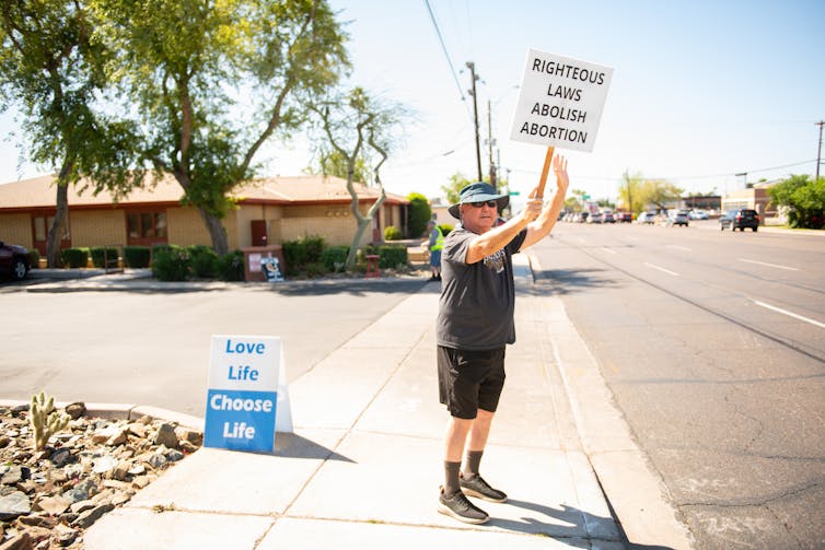 A man holds a sign that says 'righteous laws abolish abortion' and in front of a sign that says 'love life, choose life' on a quiet street.