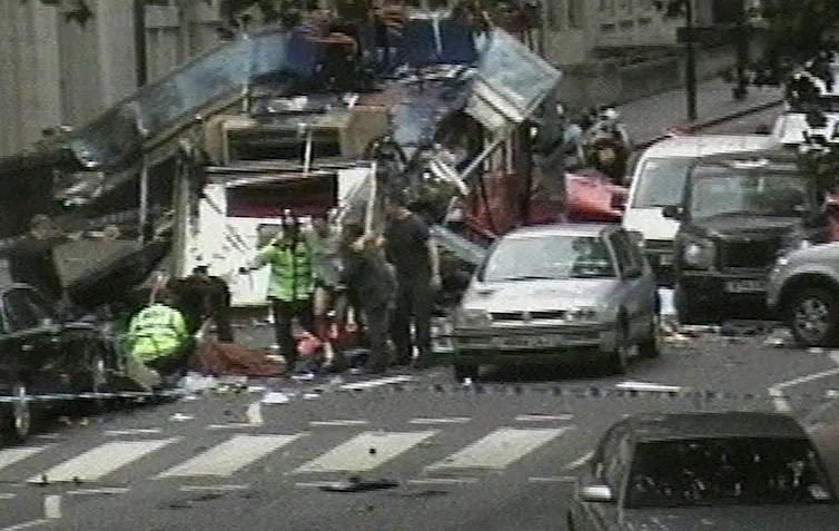 A street with the blown up wreckage of a bus, surrounded by police and behind police tape.
