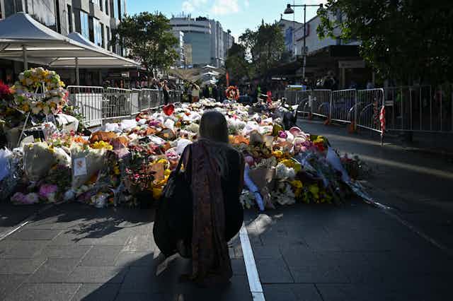 woman kneels on street looking at many flowers laid in tribute outside shopping centre where deaths occurred