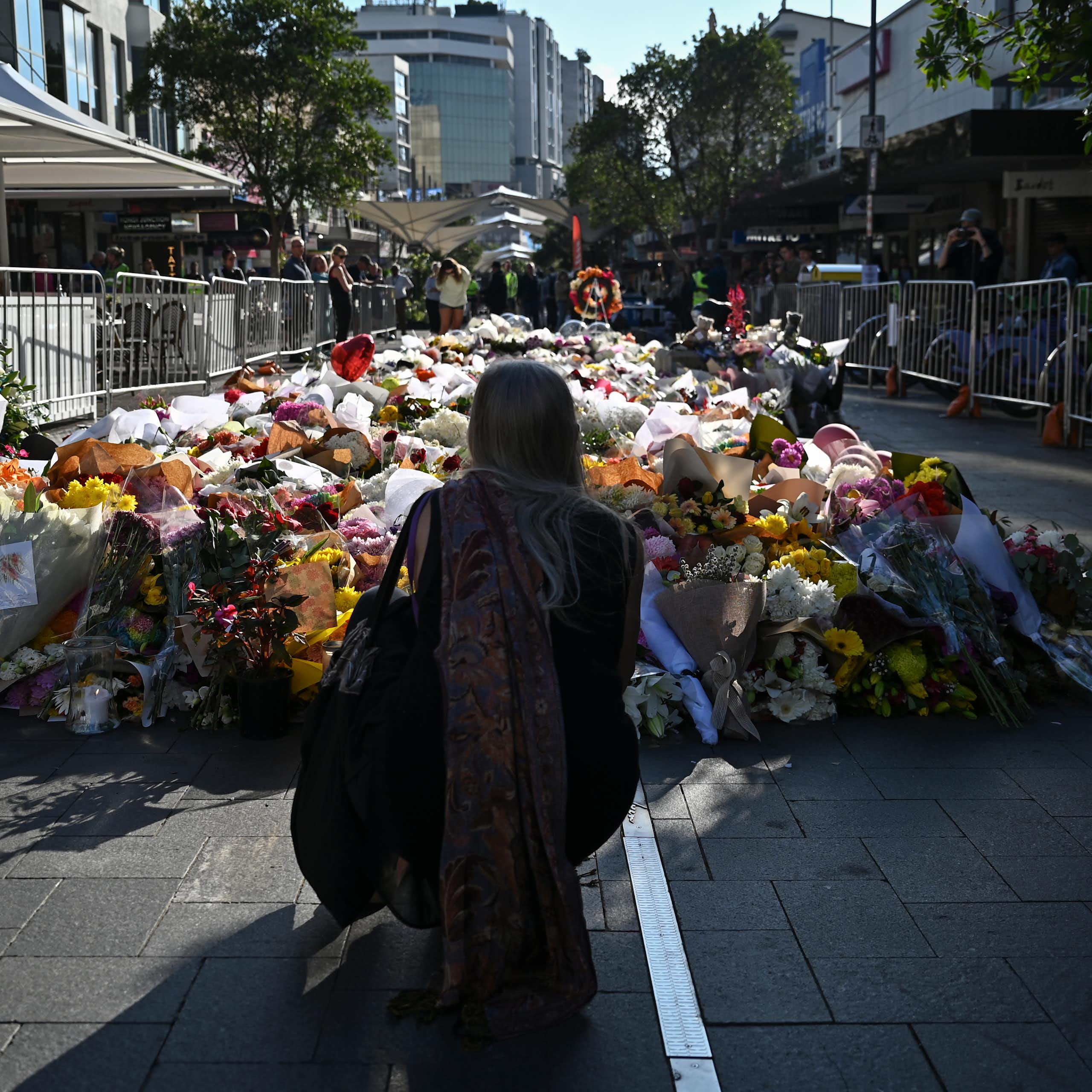 woman kneels on street looking at many flowers laid in tribute outside shopping centre where deaths occurred