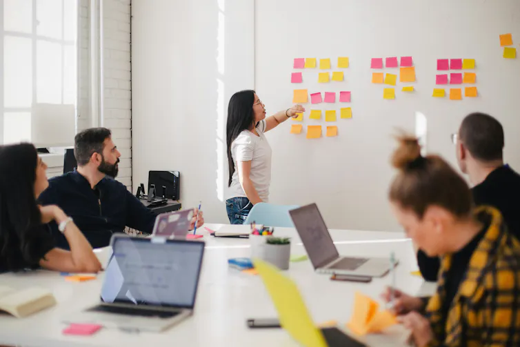 Woman places sticky notes on wall in front of a meeting of colleagues