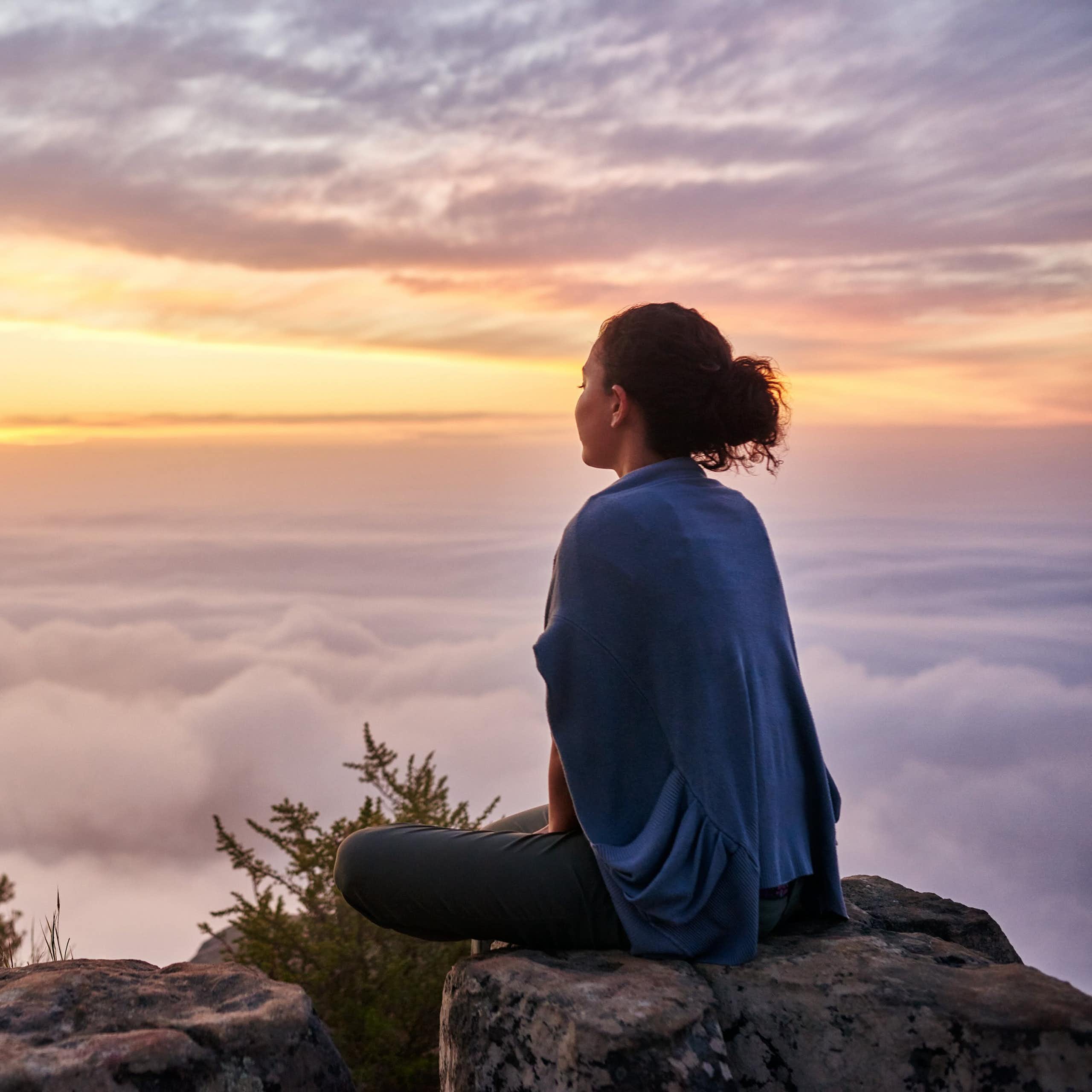 Photo of a woman sitting on top of a mountain looking out a sunrise over clouds.