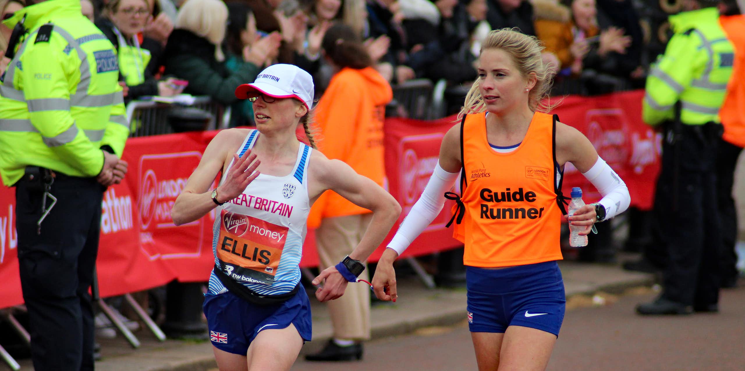 Two women running side by side, with a short tether connecting their wrists. Charlotte Ellis is wearing a race bib with her last name and a Great Britain vest, her guide runner is wearing a flourescent orange Guide Runner vest