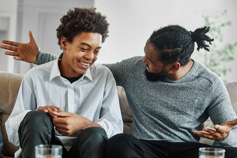 A portrait of a laughing teenage boy son talking to his father on their couch.