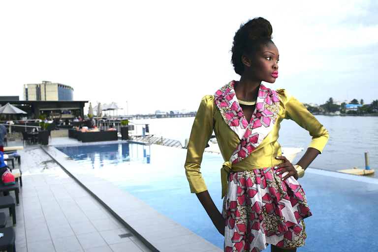 Une jeune femme se tient au bord d'une piscine, un bras sur la hanche, et pose dans une tenue d'un vert brillant avec un tissu africain rose contrastant au niveau du col et de la jupe.
