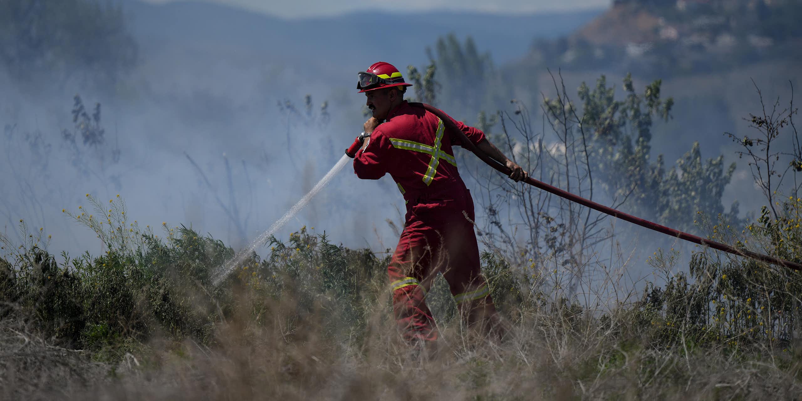 A firefighter points his hose at a grass fire with hills behind him.