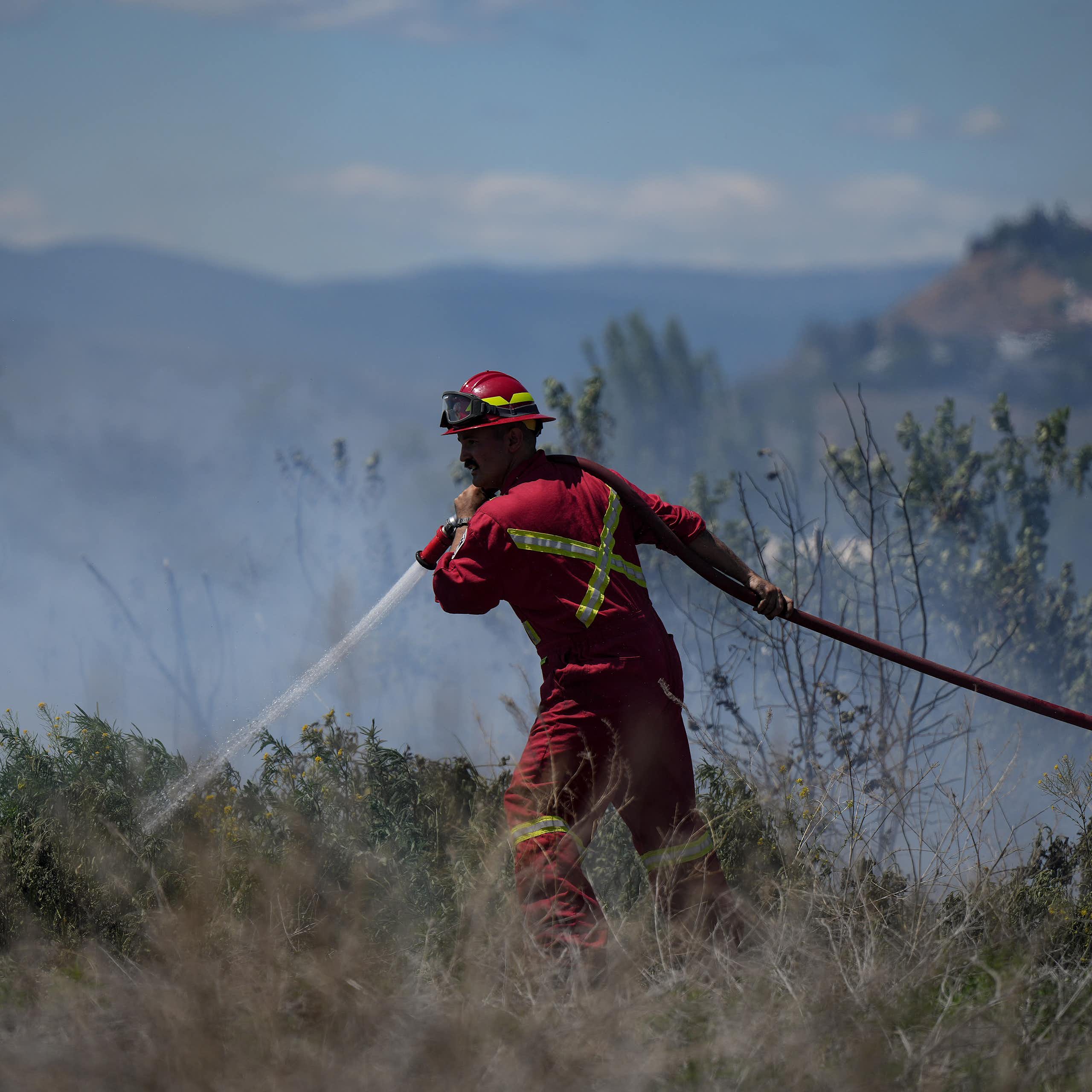 A firefighter points his hose at a grass fire with hills behind him.