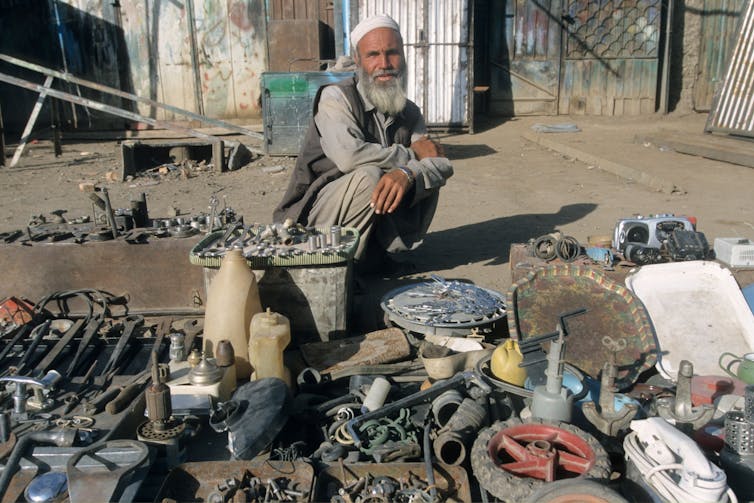 An Afghan man sitting on the floor in front are spread metal objects he is selling.