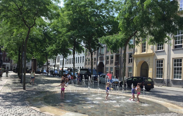 city street with trees, green leaves and shade, water fountains spouting up form the paving, kids in swimming shorts playing in the water, buildings in background