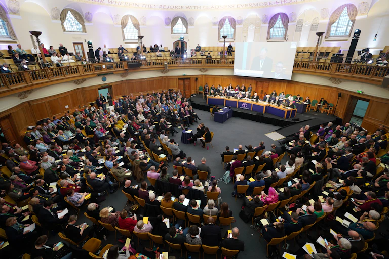 People seated in several rows arranged in the well of a circular hall, while several watch from a level above.
