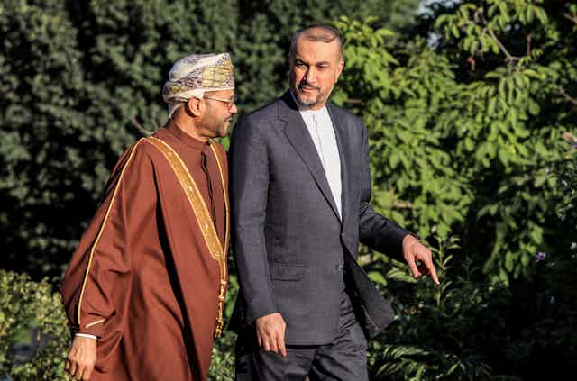 Two men, one in traditional Gulf state attire, walk in front of some trees.
