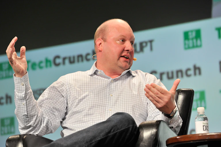 A man in a chair gestures while talking. A technology conference banner is displayed behind him.