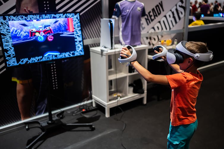 Young boy plays with a VR headset while looking at a huge computer monitor screen with both hands outstretched.