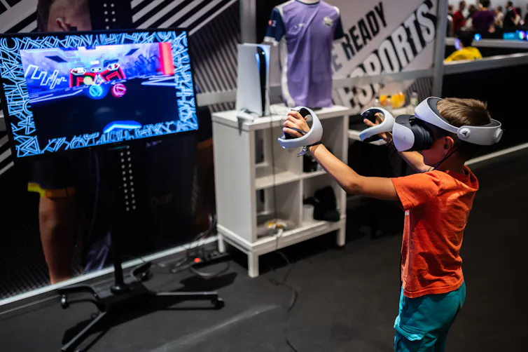 Young boy plays with a VR headset while looking at a huge computer monitor screen with both hands outstretched.