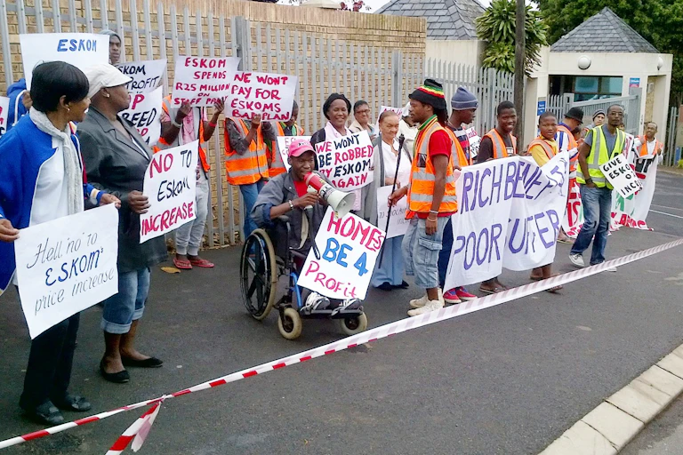 A group of people standing in a street (one in a wheelchair with a megaphone) behind barrier tape and holding placards.