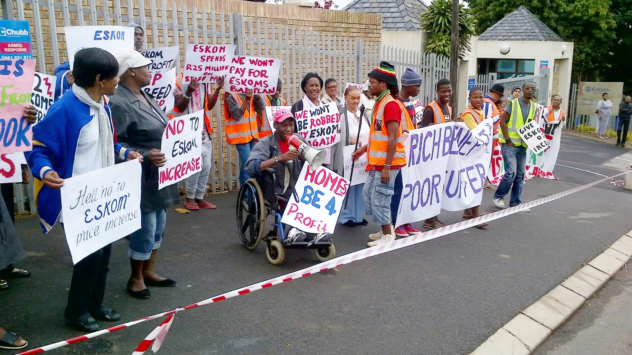 A group of people standing in a street (one in a wheelchair with a megaphone) behind barrier tape and holding placards.