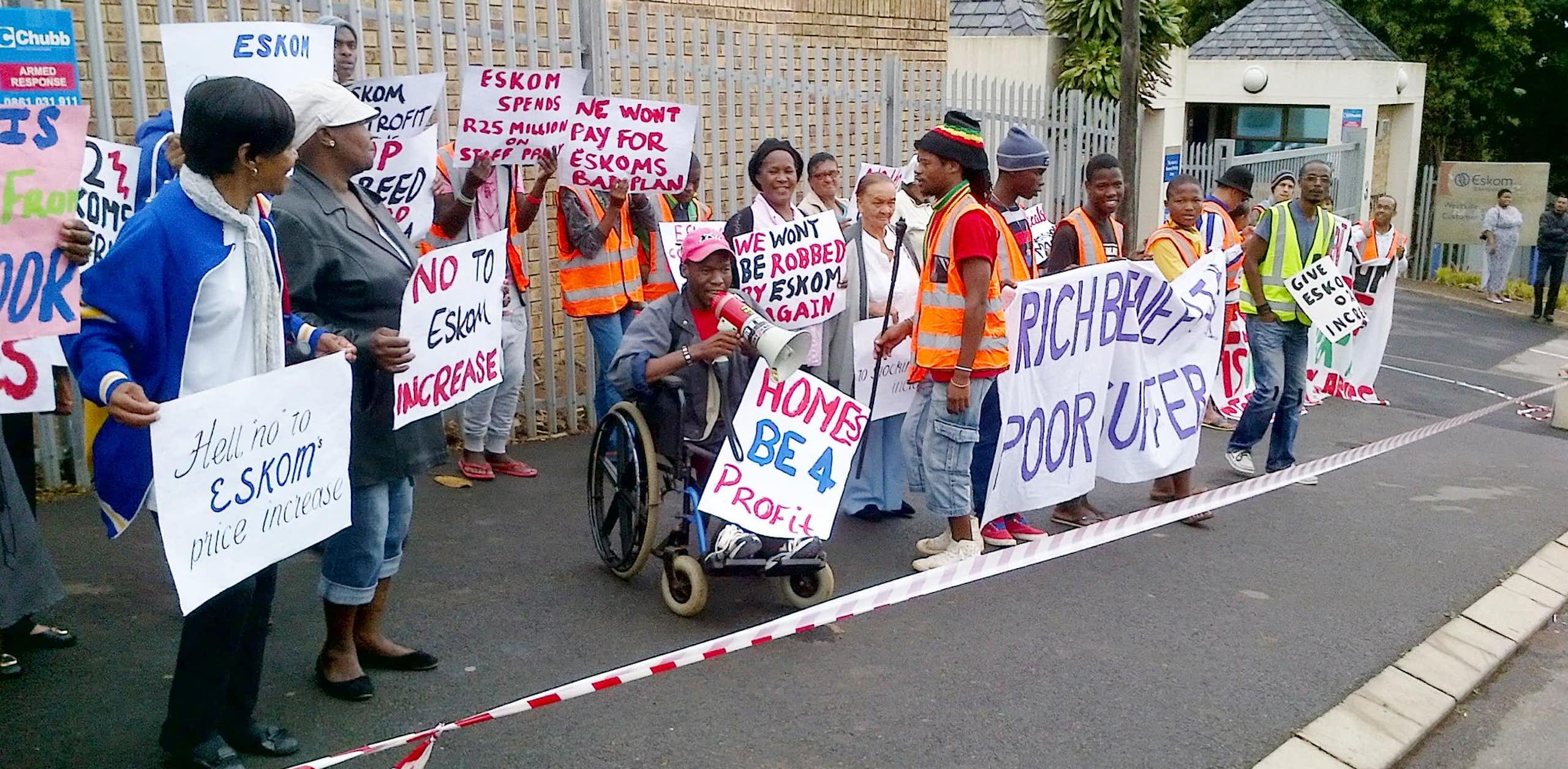 A group of people standing in a street (one in a wheelchair with a megaphone) behind barrier tape and holding placards.