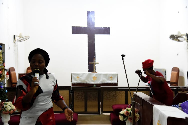 A black woman speaks into the microphone while standing next to a large crucifix placed on an altar.