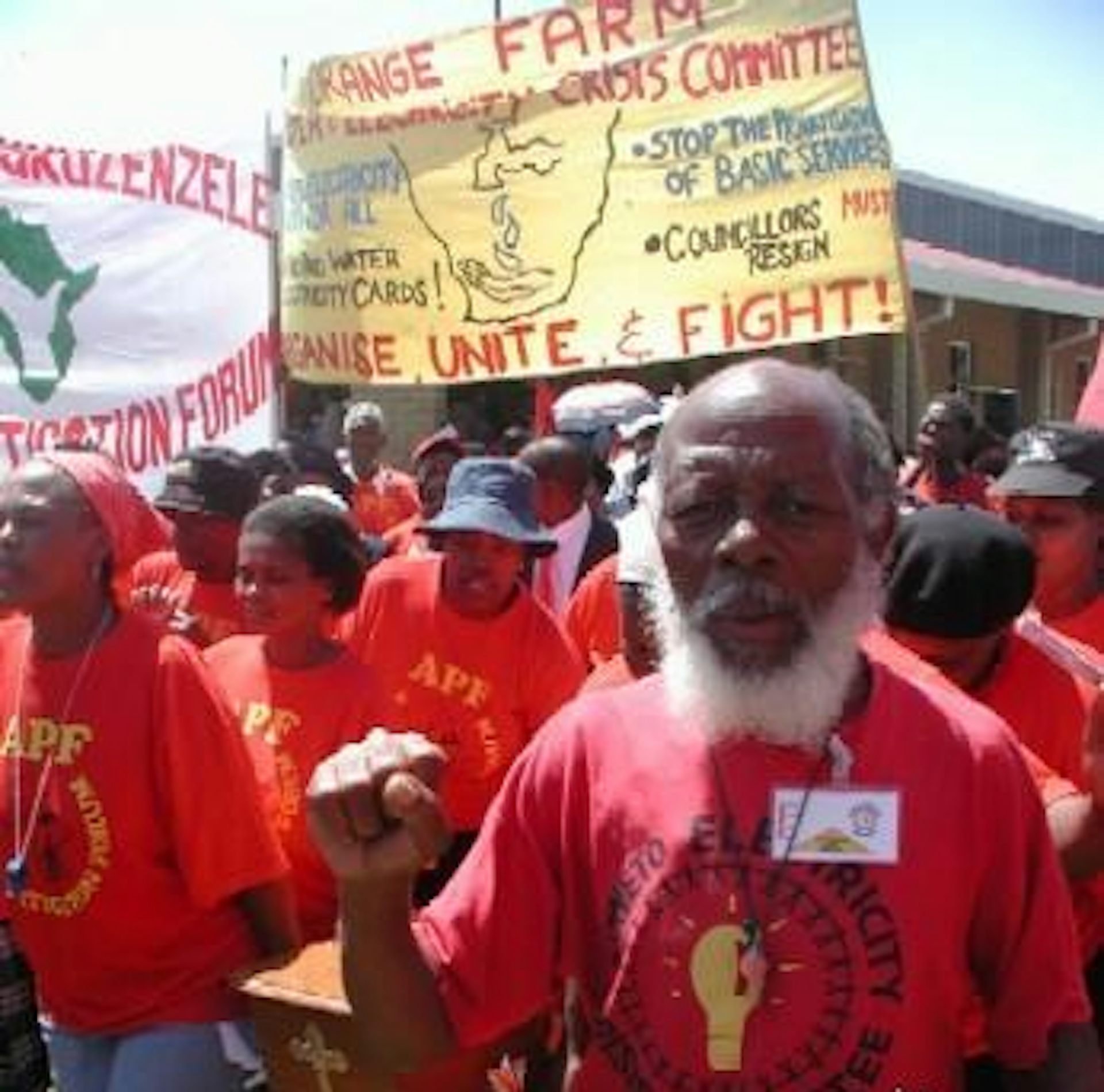 A group of people on a protest wearing red tshirts with fists raised. Banner says Organise, Unite and Fight