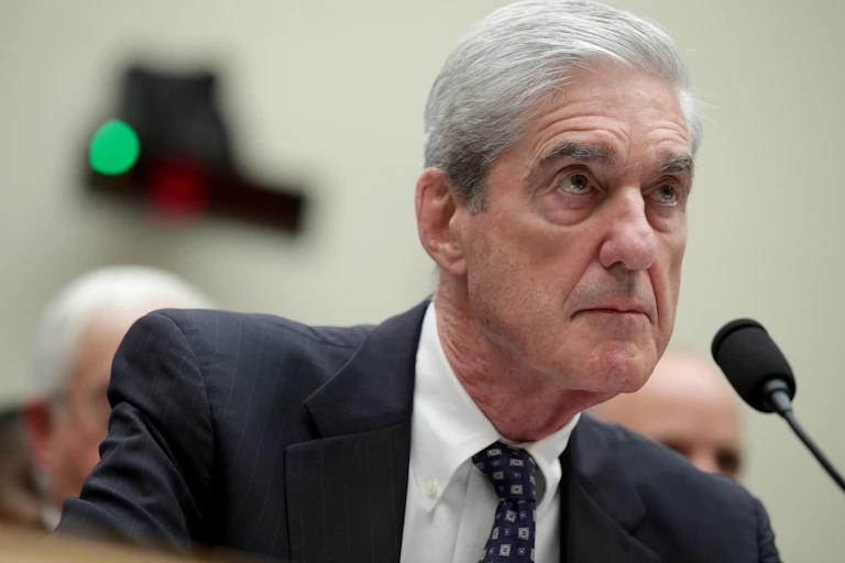 A white man with gray hair sits behind a microphone on a desk.