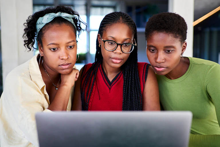 Three young Black women look at a laptop screen together.