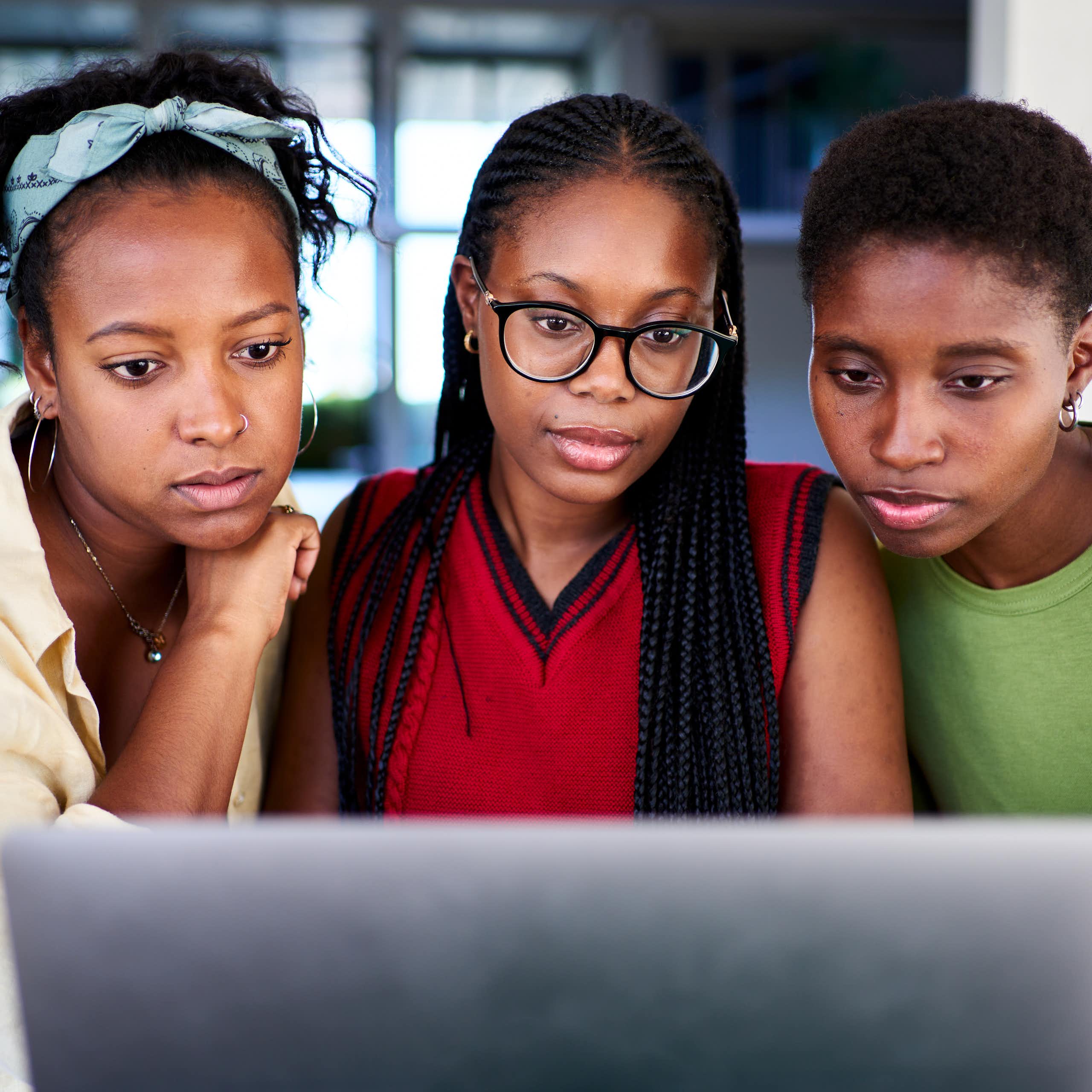 Three young Black women look at a laptop screen together.