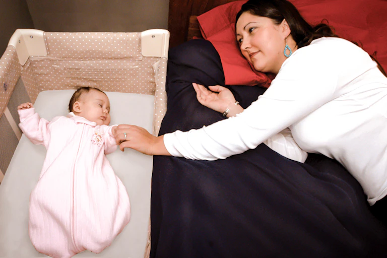 A baby sleeps in a crib wrapped in a swaddle, with the mom in her own bed next to the baby.