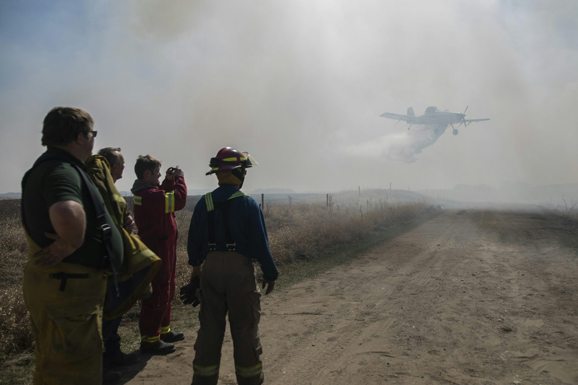 Men in firefighting equipment watch a water plane dumping water on a wildfire on the near horizon.