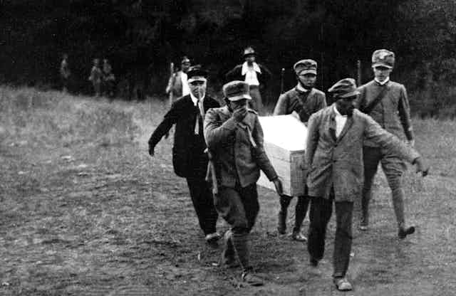 A group of uniformed men carry a coffin along a path out of woods 