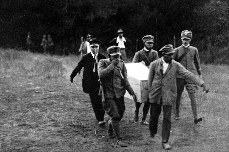 A group of uniformed men carry a coffin along a path out of woods