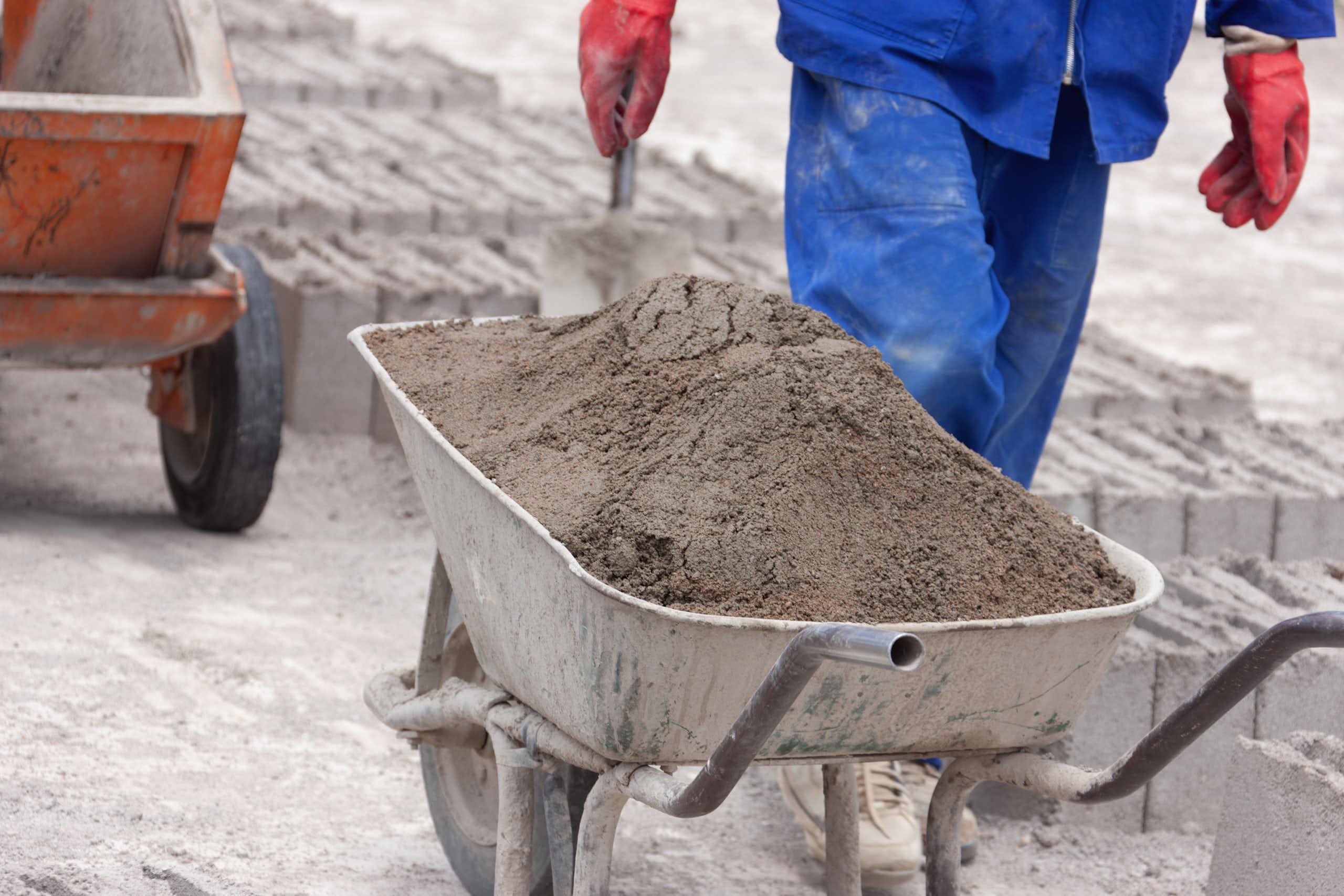 Wheelbarrow of cement and bricks in the background; person wearing work overalls and gloves walking past