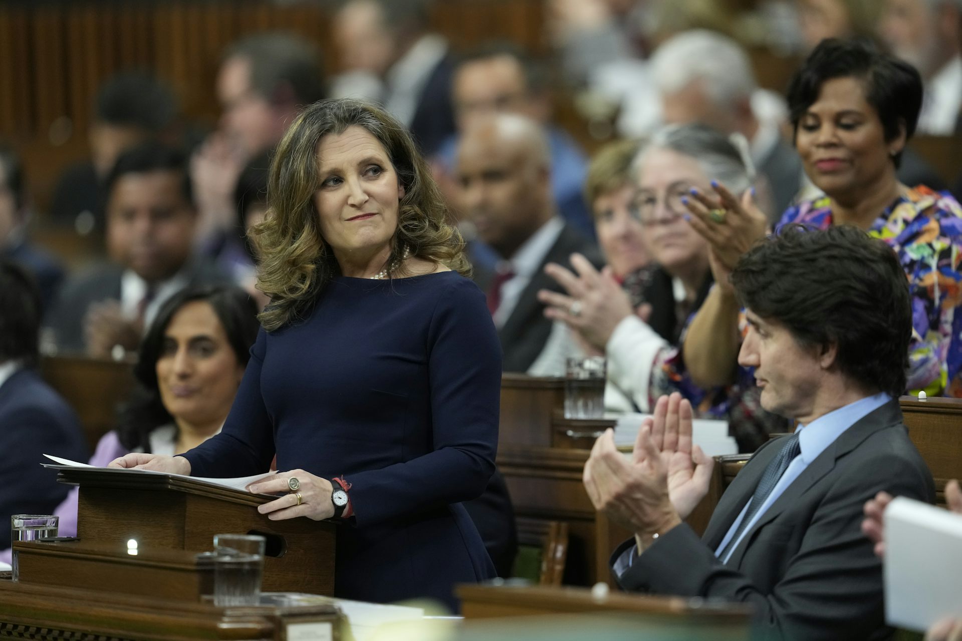 A woman in a navy dress with dark blonde hair grins while standing in the House of Commons. People around her are clapping.