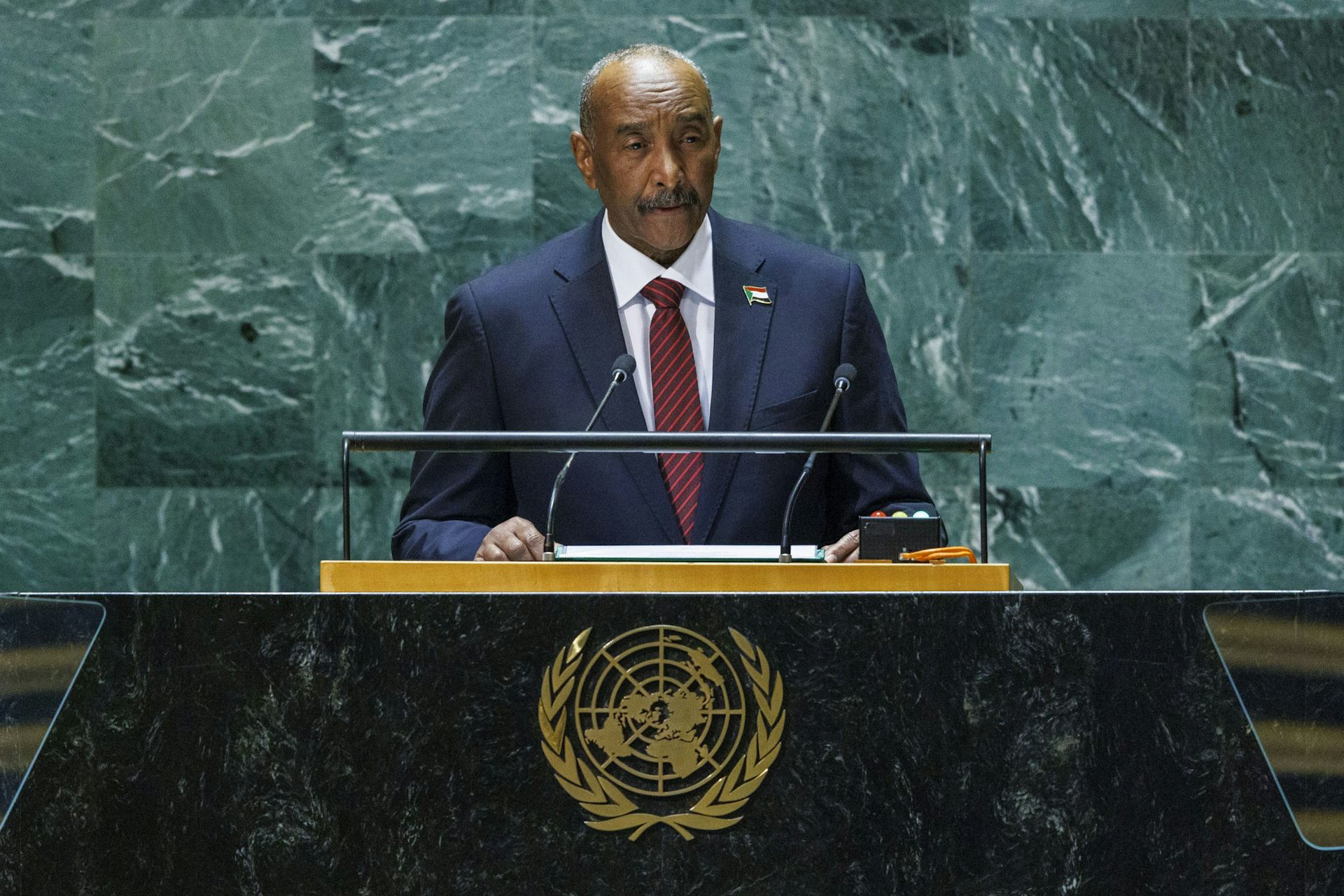 A man in a suit standing at a podium in front of a marble wall delivering a speech.