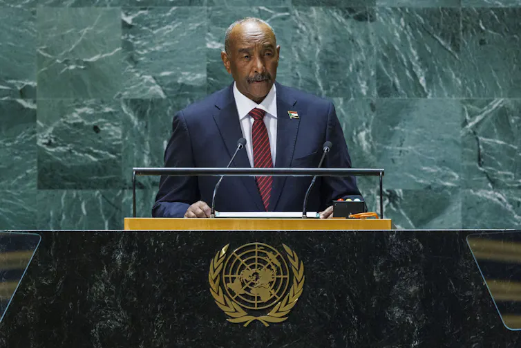 A man in a suit standing at a podium in front of a marble wall delivering a speech.