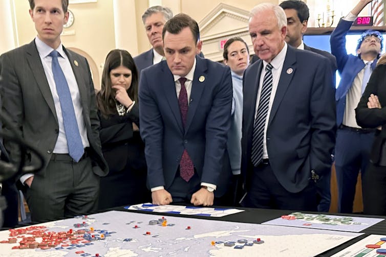 A group of people, mostly men in suits, look over a board game in an office.