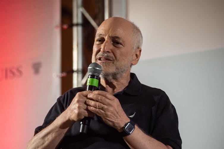 An older man in a black polo short speaks into a microphone.