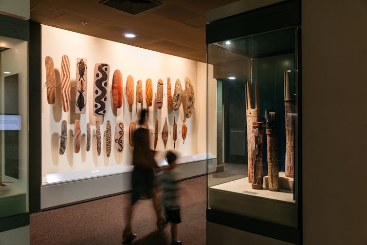 Photo of people walking past a glass case containing bark shields painted with elaborate patterns.