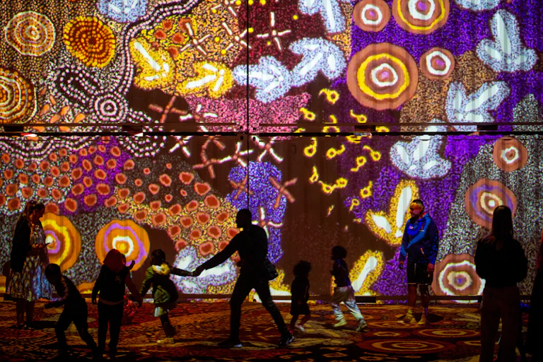 Silhouettes of people against a light installation of multicoloured Aboriginal art work