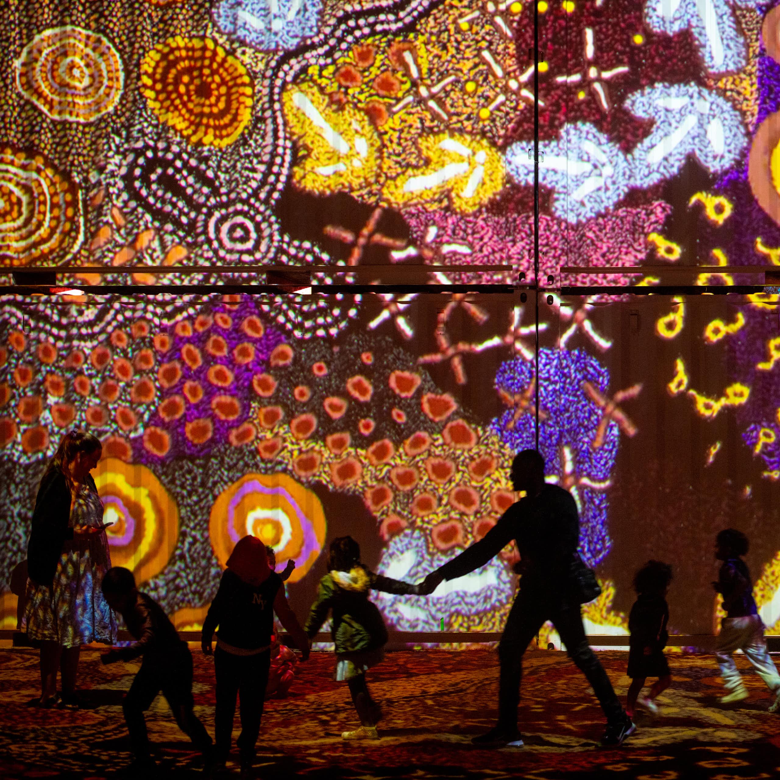 Silhouettes of people against a light installation of multicoloured Aboriginal art work