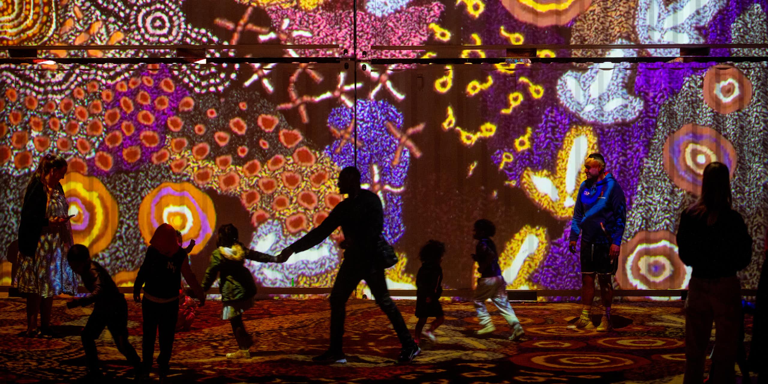 Silhouettes of people against a light installation of multicoloured Aboriginal art work