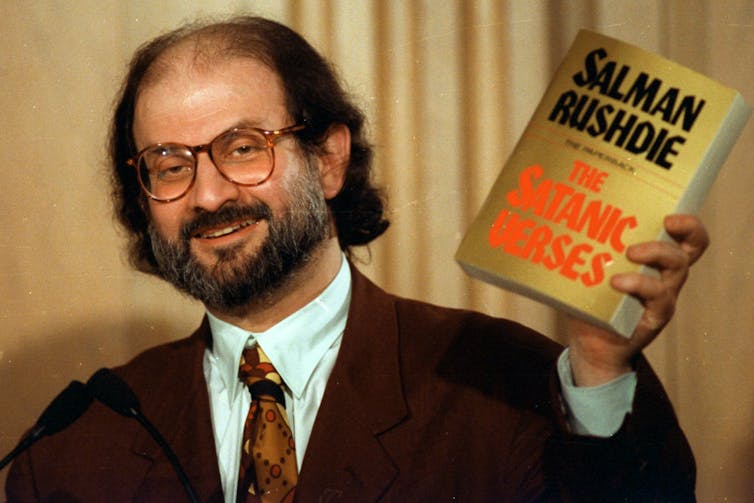 A bearded man in a suit and glasses holds up a copy of The Satanic Verses
