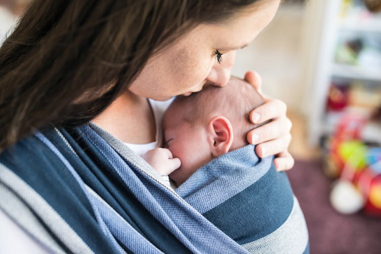 A mother kisses her baby on the head while he sleeps in a scarf.