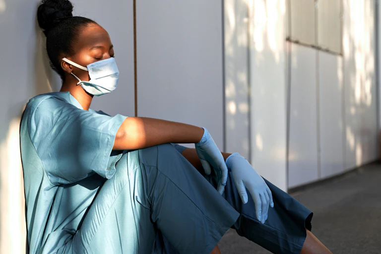 A young, black, female care worker in a face mask and gloves sits on the ground against a wall, looking exhausted