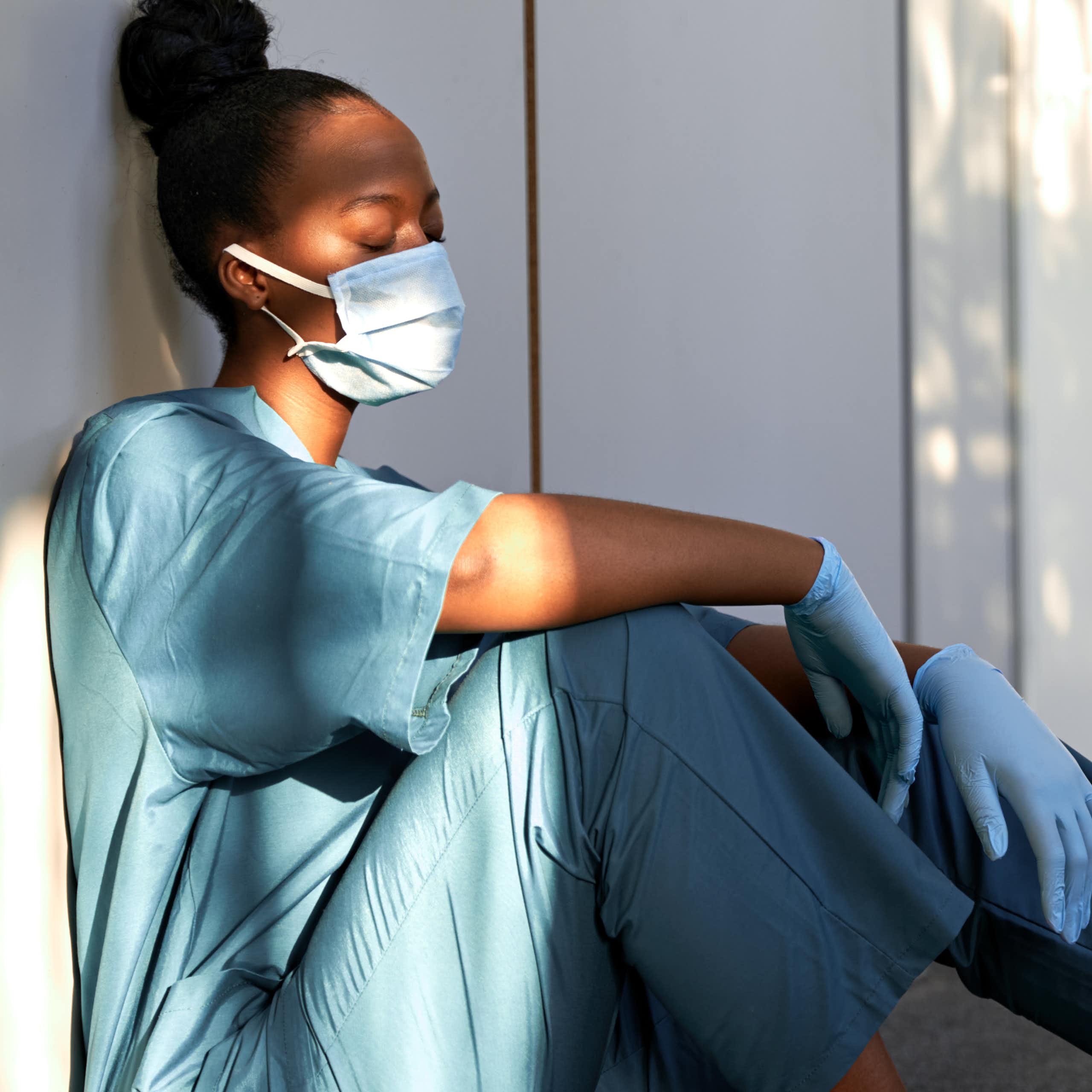 A young, black, female care worker in a face mask and gloves sits on the ground against a wall, looking exhausted