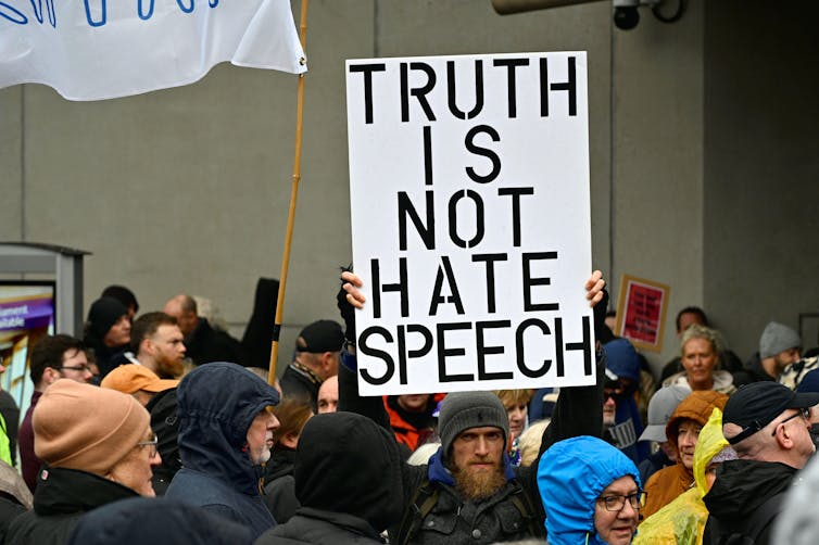 A man in a crowd holding a sign that reads 'truth is not hate speech' in large letters.